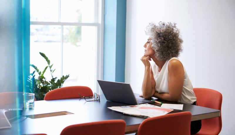 Mature woman looks out window with laptop at board room table